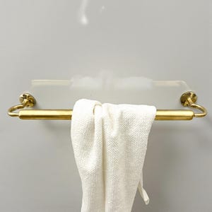 May include: A gold-colored towel bar with a white towel hanging from it. The bar is mounted on a white surface. The towel is folded over the bar. The towel bar has decorative circular end pieces.