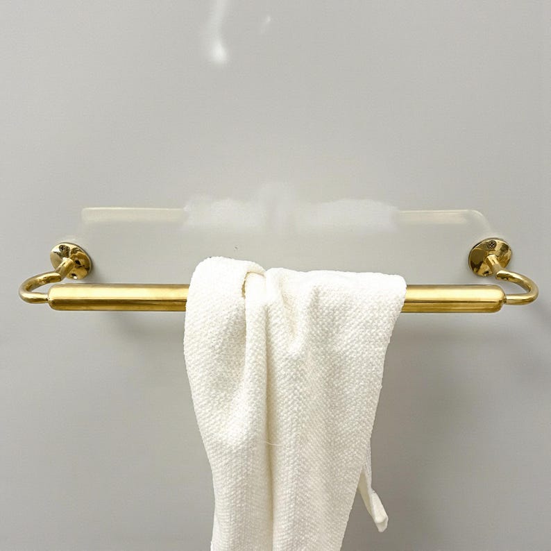 May include: A gold-colored towel bar with a white towel hanging from it. The bar is mounted on a white surface. The towel is folded over the bar. The towel bar has decorative circular end pieces.