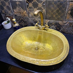 May include: An antique-style, oval-shaped, gold-colored sink with an ornate floral design around the rim. The sink has a matching gold faucet with two cross-shaped handles. The sink is set against a backdrop of decorative black and gold tiles.
