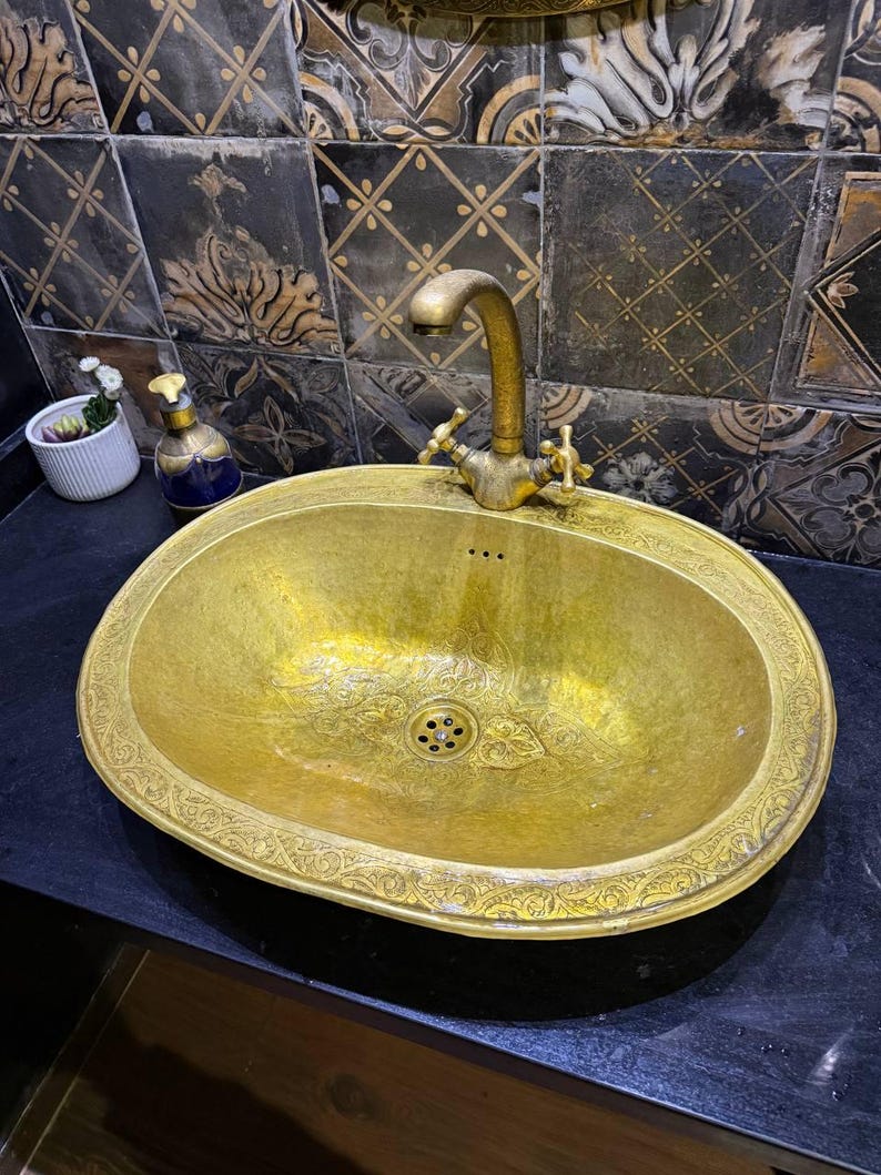 May include: An antique-style, oval-shaped, gold-colored sink with an ornate floral design around the rim. The sink has a matching gold faucet with two cross-shaped handles. The sink is set against a backdrop of decorative black and gold tiles.
