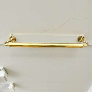 May include: A gold-colored towel bar with a cylindrical design. The bar is mounted on a white rectangular base with two circular, decorative end pieces. The bar is made of a shiny, metallic material.