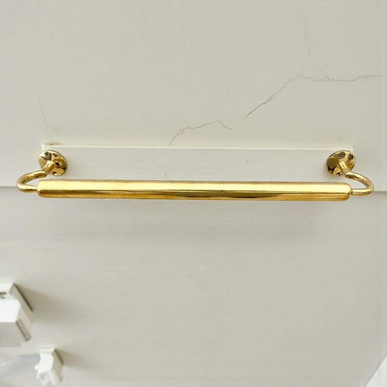 May include: A gold-colored towel bar with a cylindrical design. The bar is mounted on a white rectangular base with two circular, decorative end pieces. The bar is made of a shiny, metallic material.