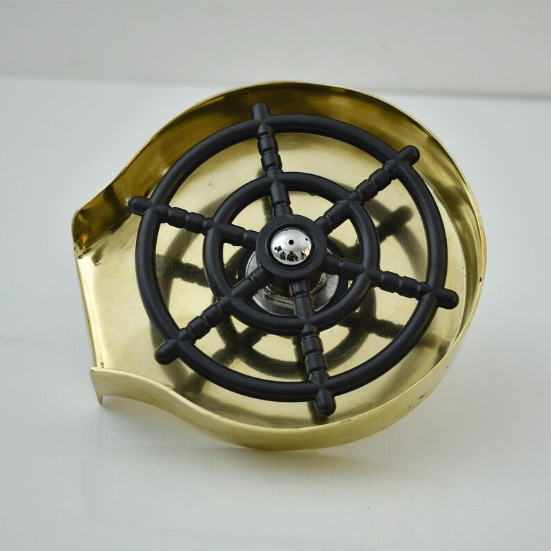 K&ouml;nnte beinhalten: Ein messingfarbener Bar-Sp&uuml;lbecken-Rinser mit einem schwarzen Schiffsraddesign. Der Rinser hat eine runde Form mit einem kleinen, erh&ouml;hten Rand und einer zentralen D&uuml;se. Das Steuerrad ist aus schwarzem Kunststoff.