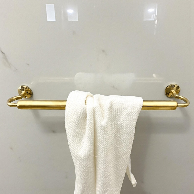 May include: A gold-colored towel bar with a white towel hanging from it. The bar is mounted on a white marble wall. The towel is folded over the bar.