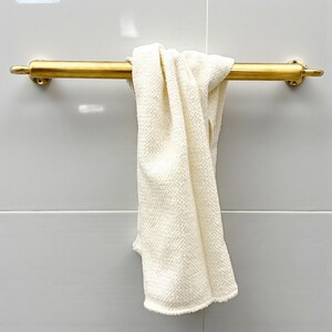 May include: A gold-colored towel bar with a cream-colored towel hanging from it. The towel bar is mounted on a light gray tiled wall. The towel is folded over the bar.