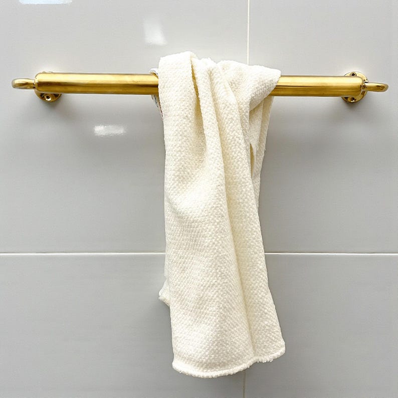 May include: A gold-colored towel bar with a cream-colored towel hanging from it. The towel bar is mounted on a light gray tiled wall. The towel is folded over the bar.