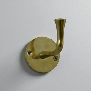 May include: A brass wall hook with a curved arm and a round base. The hook is mounted with two screws and has a polished finish. The hook is designed for hanging items and is a decorative hardware piece.