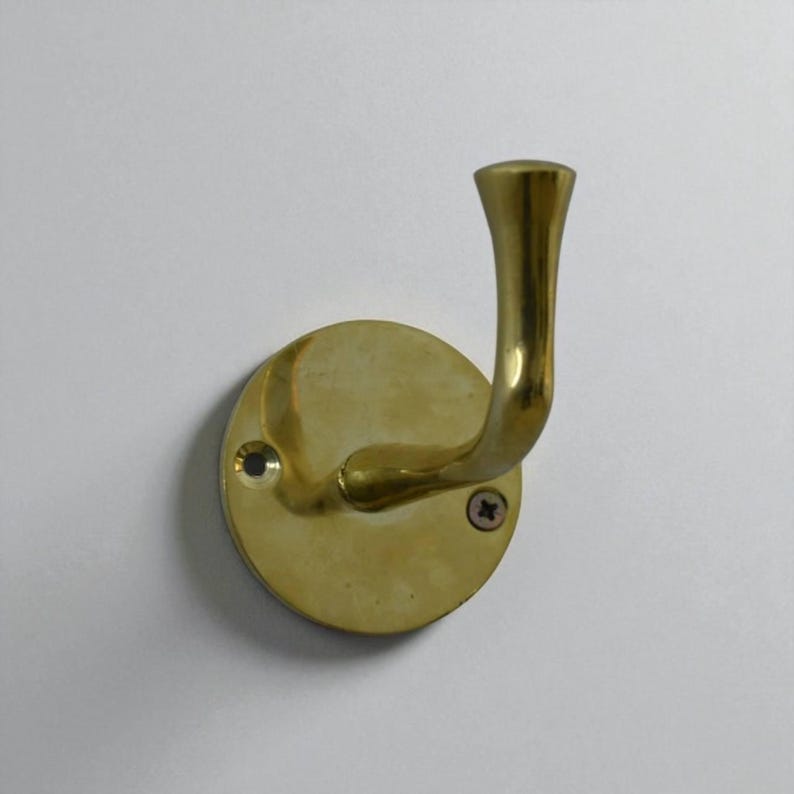 May include: A brass wall hook with a curved arm and a round base. The hook is mounted with two screws and has a polished finish. The hook is designed for hanging items and is a decorative hardware piece.