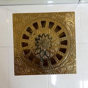 Könnte beinhalten: Ein quadratischer, goldfarbener Abdeckrost mit einem verzierten, gravierten Blumenmuster. Die Mitte weist ein kreisförmiges Design mit einem Sternmuster auf. Der Abdeckrost besteht aus Metall und ist wahrscheinlich für eine Dusche oder einen Bodenablauf.