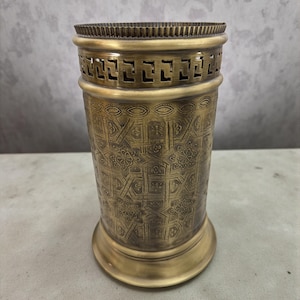 May include: A cylindrical brass vase with intricate geometric and floral patterns. The vase has a wide base and a decorative band with rectangular cutouts near the top. The overall design is antique and ornate.