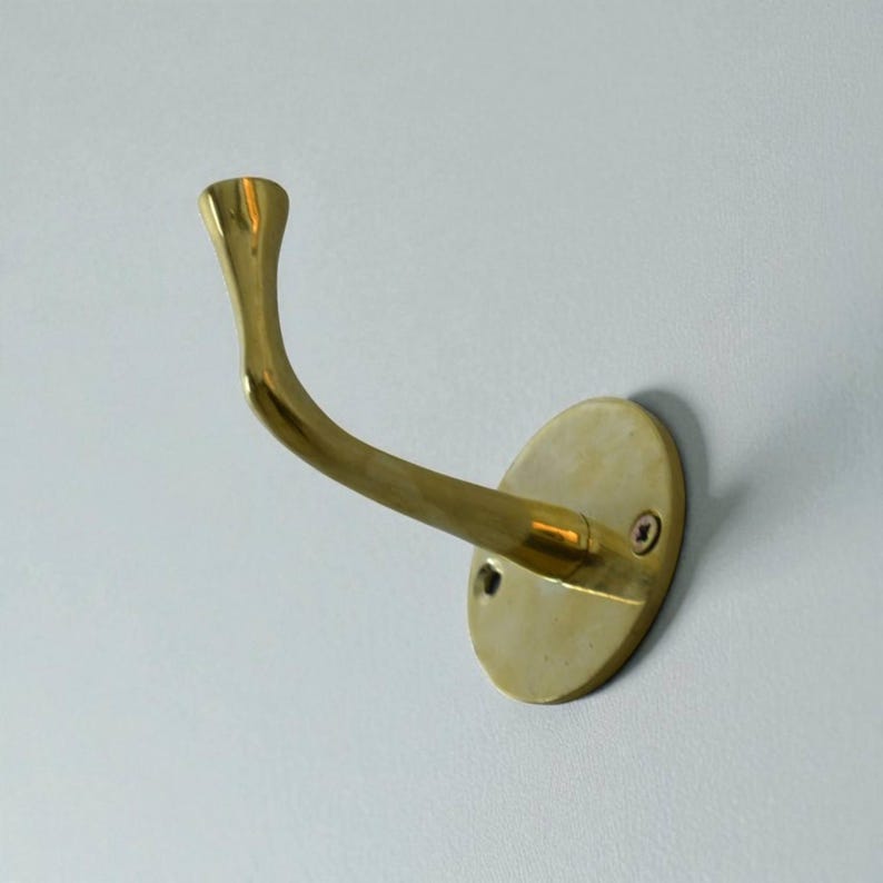 May include: A brass wall hook with an oval base and a curved arm. The hook is a warm gold color and is mounted on a light gray wall. The hook is designed to hold coats, hats, or other items.