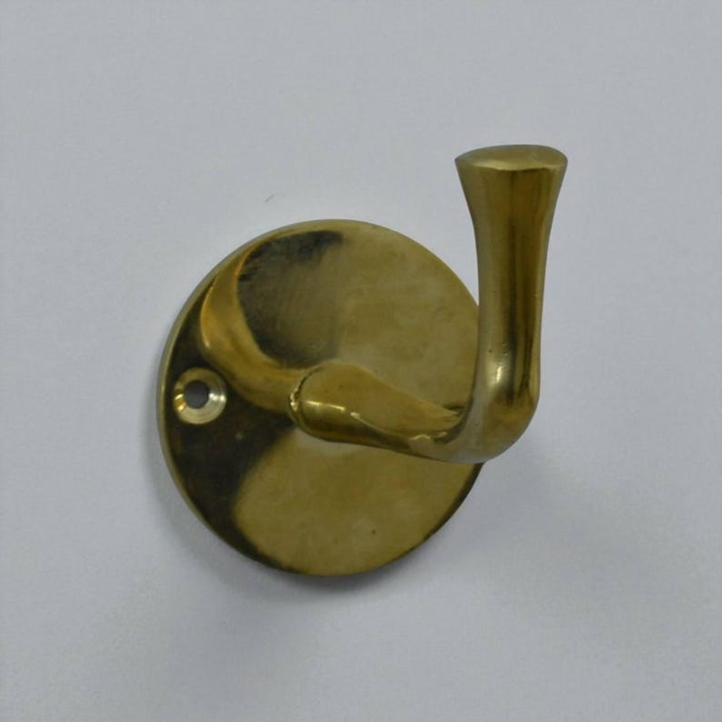 May include: A brass-colored wall hook with a curved design. The hook is mounted on a circular base with a single screw hole. The hook has a smooth, polished finish and is designed for hanging items.