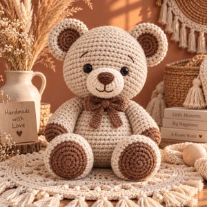 May include: A crocheted teddy bear featuring a light beige body with dark brown accents on the ears, paws, and a small bow tie at the neck. The bear has black plastic eyes, a dark brown stitched nose, and a simple stitched mouth on its face.