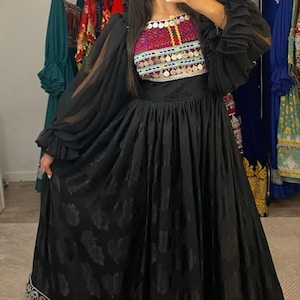 Puede incluir: Un vestido negro con falda estampada y mangas largas y transparentes. El corpiño presenta bordados coloridos y detalles metálicos. El vestido tiene un dobladillo detallado, probablemente una prenda formal o cultural.