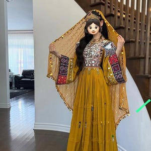 Könnte beinhalten: Ein senfgelbes traditionelles Kleid mit aufwendiger Stickerei und Spiegelverzierungen. Das Kleid hat lange Ärmel mit detaillierten Mustern und eine passende Kopfbedeckung. Goldmünzen schmücken die Ränder der Kopfbedeckung.