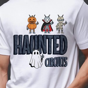 May include: White t-shirt featuring a Halloween-themed design. The shirt displays three cartoon characters: a pumpkin, a vampire, and a mummy, along with a ghost. The text "HAUNTED CIRCUITS" is printed below the characters in a blue and black font.