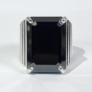 Puede incluir: Un anillo con una gran piedra negra rectangular y una banda plateada. El anillo presenta una piedra oscura prominente engastada en un engaste plateado con líneas verticales en los lados. El anillo se muestra sobre una superficie blanca.