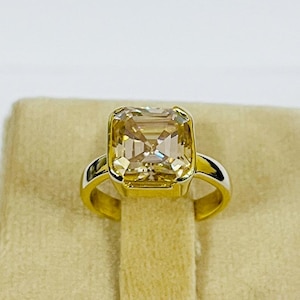 Może przedstawiać: Złoty pierścionek z dużą, kwadratowo oszlifowaną, jasnożółtą szlachetnym kamieniem. Kamień osadzony jest w złotej obrączce o prostym, eleganckim wzornictwie. Pierścionek jest prezentowany na beżowej powierzchni.