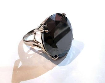May include: A silver ring featuring a large, faceted black gemstone. The gemstone is oval-shaped and catches the light, displaying multiple facets. The ring has a simple, elegant design with a silver band and setting.