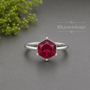 Könnte beinhalten: Ein silberner Ring mit einem sechseckigen, tiefroten Edelstein. Der Edelstein ist der Mittelpunkt, gefasst in einem silbernen Band mit kleinen, runden Akzenten an jedem Punkt des Sechsecks. Text: "Rkjewelsdesign".