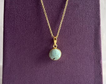 Peut inclure: Un collier doré avec un petit pendentif rond en turquoise. Le pendentif est bleu-vert clair avec des veines gris foncé. Le collier est présenté sur une surface violette texturée.