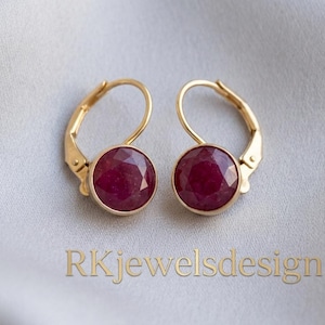 May include: A pair of gold leverback earrings featuring round, faceted, deep red rubies. The metal settings are polished gold, and the text RKjewelsdesign is printed in a light brown font below the earrings.