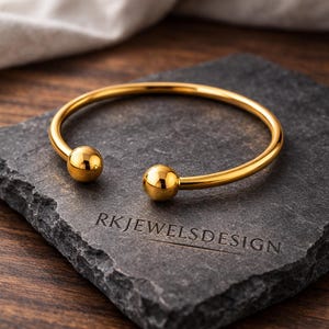 May include: A gold-colored bangle bracelet with a smooth, rounded design. The bracelet features a spherical accent at each end. The bracelet is displayed on a dark gray stone surface with the text "RK JEWELSDESIGN" engraved on it.