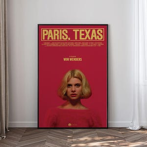 Puede incluir: Un póster de película enmarcado de "Paris, Texas" con un fondo rojo. El título está en letras doradas en la parte superior, con el nombre del director debajo. Una mujer con cabello rubio y un suéter rojo está centrada en el póster.