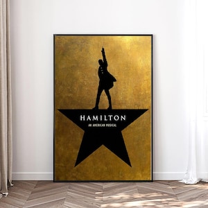 May include: A framed poster featuring the silhouette of a person with an arm raised, standing on a black star. The poster has the word "HAMILTON" above "AN AMERICAN MUSICAL" on a gold background.