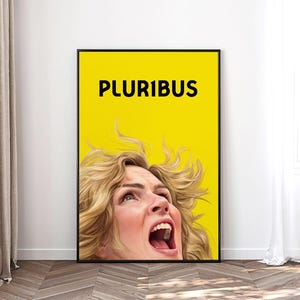 May include: A framed art print featuring a woman with blonde hair, her mouth open in a scream, set against a bright yellow background. The word "PLURIBUS" is printed in black at the top. The artwork is displayed in a room with a white wall and a curtain.