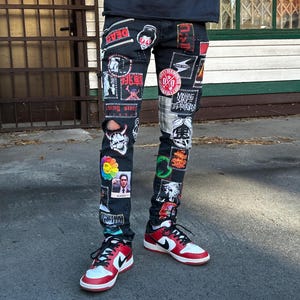 May include: Black denim jeans covered in patches with various designs and text. The patches include images of skulls, flowers, and Japanese text. The jeans are paired with red, white, and black sneakers.