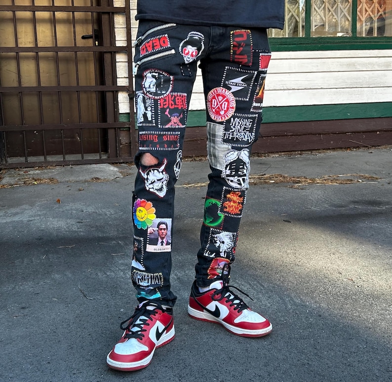May include: Black denim jeans covered in patches with various designs and text. The patches include images of skulls, flowers, and Japanese text. The jeans are paired with red, white, and black sneakers.
