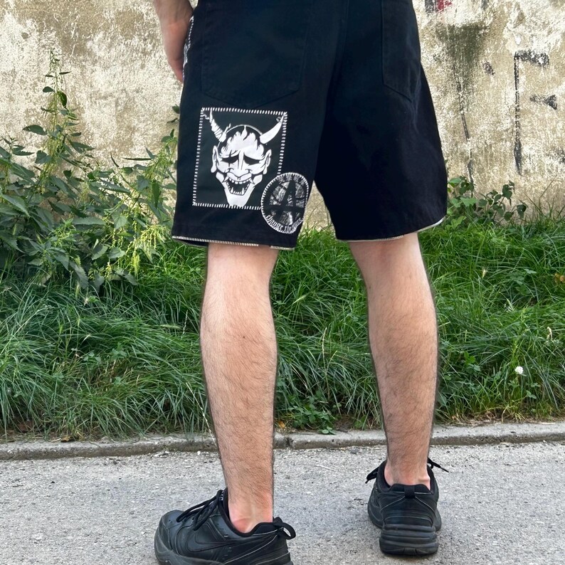 Puede incluir: Pantalones cortos de mezclilla negros con costuras blancas y un parche con borde blanco con una m&aacute;scara de demonio y un gr&aacute;fico circular. Los pantalones cortos tienen un dise&ntilde;o cl&aacute;sico de cinco bolsillos.