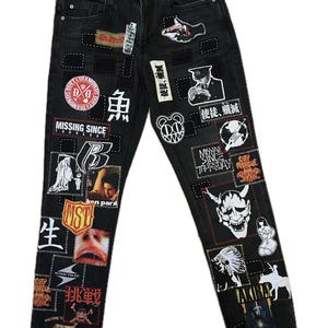 Può includere: Jeans neri in denim con varie toppe con testo e grafica. Le toppe includono le parole "Missing Since Thursday", "City Morgue" e caratteri giapponesi. I jeans hanno una chiusura con bottone e cerniera.