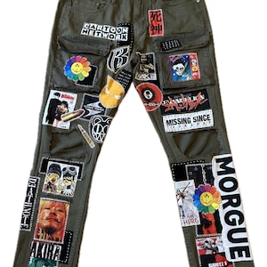 May include: Olive green denim pants with multiple patches featuring cartoon characters, Japanese text, and the words "MORGUE" and "MISSING SINCE THURSDAY." The pants have a distressed look with visible stitching and pockets.