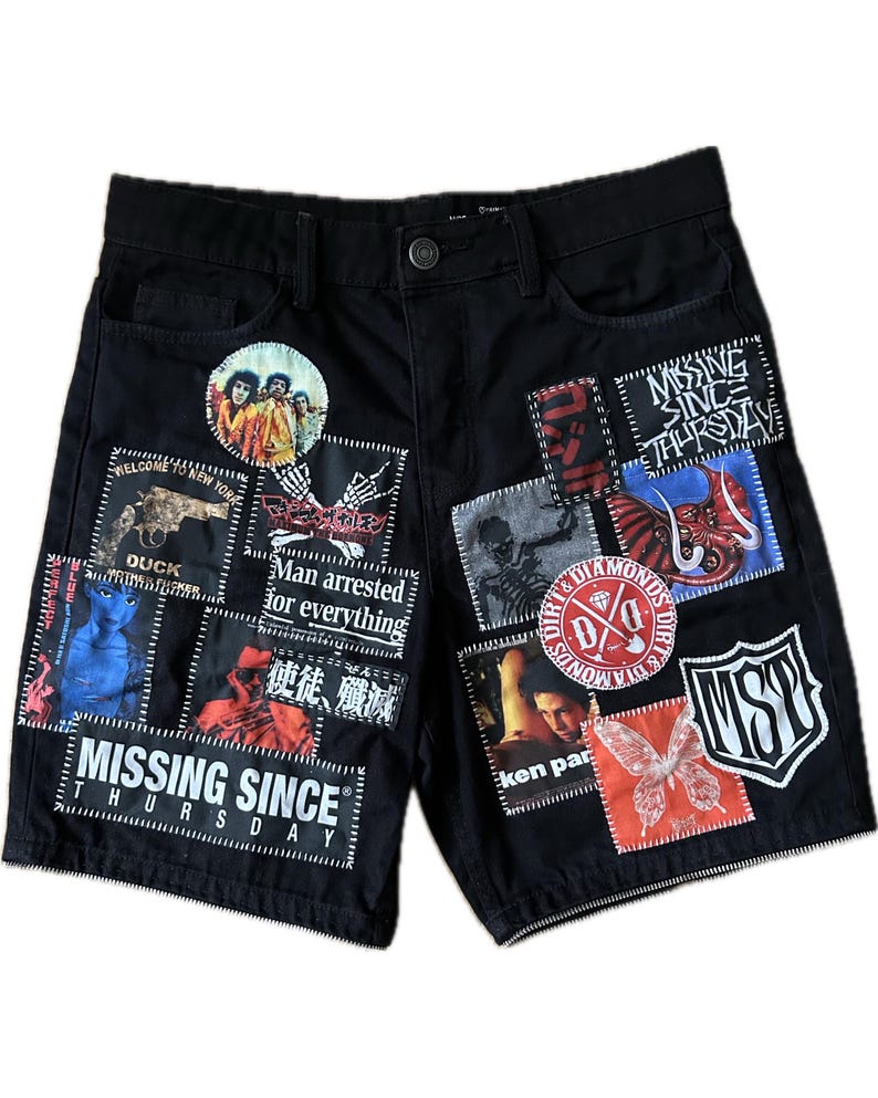 Puede incluir: Pantalones cortos de mezclilla negros con m&uacute;ltiples parches con varios gr&aacute;ficos y texto. Los parches incluyen una pistola, un esqueleto, una foto de banda y el texto "MISSING SINCE THURSDAY". Los pantalones cortos tienen cierre de bot&oacute;n y bolsillos.