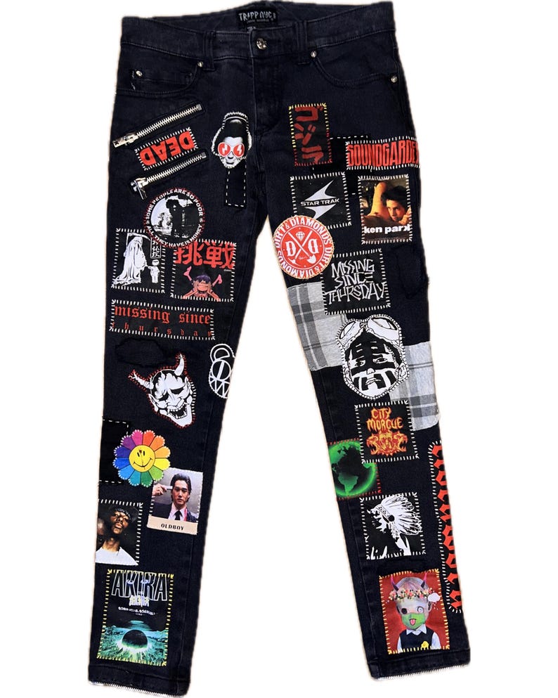May include: Black denim jeans adorned with numerous patches featuring text and graphics. Patches include the words "DEAD", "missing since", "OLDBOY", and "AKIRA". The jeans have a button and zipper closure.