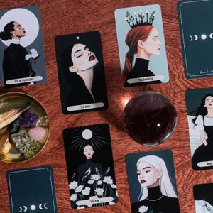 Puede incluir: Un conjunto de cartas de tarot con ilustraciones de mujeres y elementos celestiales. Las cartas son negras con texto e imágenes en blanco, incluyendo "Ace of Pentacles", "The Star", "The Empress" y "The Sun". Una bola de cristal transparente y un pequeño cuenco con cristales también están presentes.
