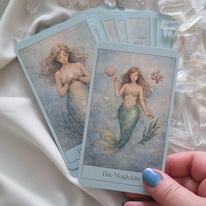 May include: A deck of tarot cards featuring mermaid illustrations. The cards have a light blue border and depict mermaids in an underwater setting with seashells and coral. One card is labeled "The Magician".