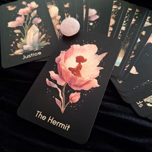 Peut inclure: Un jeu de cartes de tarot avec des illustrations florales et les mots "Justice" et "The Hermit". Les cartes présentent un fond sombre avec des fleurs roses et blanches. Un cristal rose est posé sur les cartes.