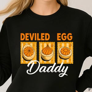 Puede incluir: Sudadera negra con el texto "DEVILED EGG Daddy" en naranja y blanco. El diseño incluye tres imágenes estilo sello postal de huevos rellenos con relleno naranja sobre un fondo a rayas amarillas y blancas.
