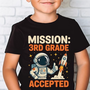 May include: Black t-shirt with the text "MISSION: 3RD GRADE ACCEPTED." The graphic features an astronaut giving a thumbs up next to a rocket blasting off, with stars in the background. The design is in shades of white, orange, and yellow.