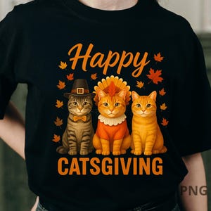 May include: Black t-shirt with the words "Happy CATSGIVING" in orange script. The graphic features three cartoon cats dressed in Thanksgiving attire, surrounded by orange maple leaves. One cat wears a pilgrim hat, another a turkey costume.