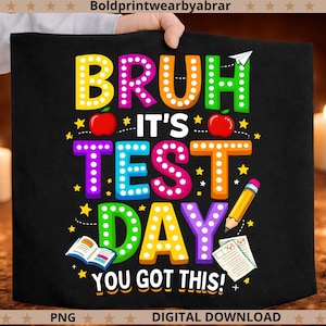 May include: A black t-shirt featuring the colourful text BRUH IT'S TEST DAY YOU GOT THIS. The design includes illustrations of two red apples, a yellow pencil, an open book, a white paper aeroplane, and a white test paper with checkmarks.