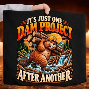 May include: A black fabric item featuring a graphic of a brown beaver wearing a yellow hard hat and carrying a bundle of logs. The text reads IT'S JUST ONE DAM PROJECT AFTER ANOTHER in orange and cream lettering against a forest and river scene.