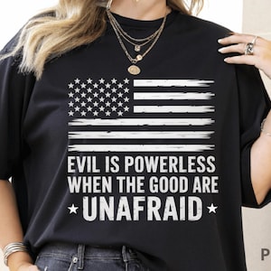 May include: Black t-shirt featuring a distressed American flag design in white. The shirt includes the text "EVIL IS POWERLESS WHEN THE GOOD ARE *UNAFAID*" in white, with star accents. A casual, patriotic apparel item.
