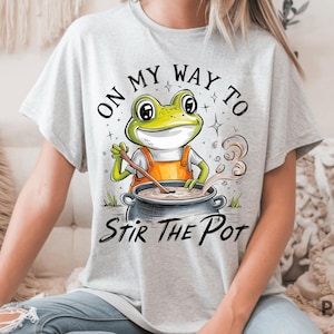 May include: A light gray t-shirt featuring a cartoon frog wearing an orange apron, stirring a pot. The text "ON MY WAY TO STIR THE POT" is arched above and below the frog illustration. The frog is green and yellow.