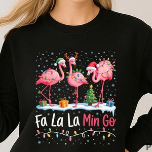 Può includere: Felpa nera con un design a tema natalizio. Presenta tre fenicotteri rosa adornati con luci natalizie e cappelli di Babbo Natale. Il testo "Fa La La Mingo" è scritto in bianco e rosa, con una fila di luci colorate sotto. Sono inclusi anche regali e un albero di Natale.