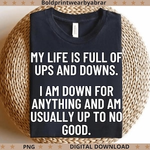 Könnte beinhalten: Ein marineblaues T-Shirt mit dem Text "MY LIFE IS FULL OF UPS AND DOWNS. I AM DOWN FOR ANYTHING AND AM USUALLY UP TO NO GOOD." Das Shirt wird auf einer gewebten, kreisförmigen Oberfläche präsentiert.