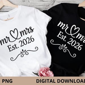 Puede incluir: Dos camisetas, una blanca y otra negra, colgadas de perchas de madera. Ambas camisetas presentan el texto "mr & mrs Est. 2026" en una fuente decorativa con un símbolo de corazón. Una flor rosa en la parte inferior.